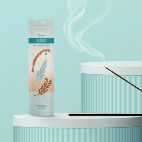 A pale blue-grey package of incense sticks that reads Flore Guidance with an image of a sweetgrass braid, sage leaves and a cedar bough. The package stands upright on a podium next to a burning incense stick against a pale aqua background.