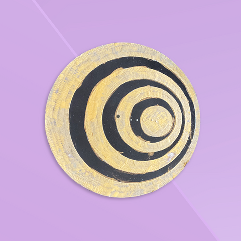 Overhead view of a round flat disk incense burner with a gold and black swirl design against a rosy pink background. 