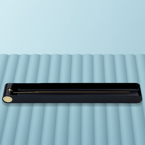 A long black metal incense burner featuring a brass knob at one end that is used to adjust the angle of an incense stick. The burner contains one stick of incense in a horizontal position and is sitting on blue-grey background. Designed and fabricated by Toronto CNC machine shop Rocket Surgery Solutions. 