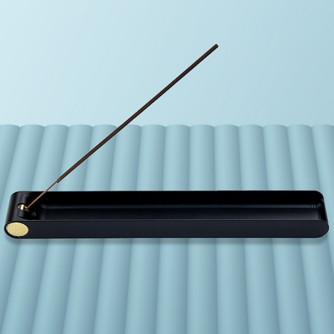 A long black metal incense burner featuring a brass knob at one end that is used to adjust the angle of an incense stick. The burner contains one stick of incense in a 45 degree angle and is sitting on blue-grey background. Designed and fabricated by Toronto CNC machine shop Rocket Surgery Solutions. 