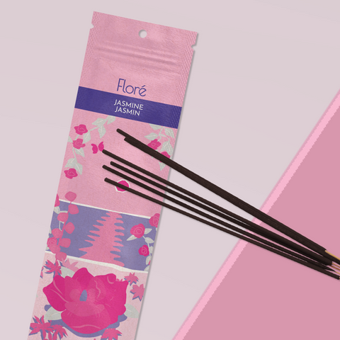 A light pink package of incense sticks that reads Flore Jasmine with an image of dark pink flowers framing a blue lake with a pink sun in the sky. The package lies with a bundle of incense sticks on a pale rose background.