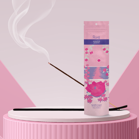 A light pink package of incense sticks that reads Flore Jasmine with an image of dark pink flowers framing a blue lake with a pink sun in the sky. The package stands upright on a podium next to a burning incense stick against a pale rose background.