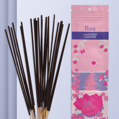 A light pink package of incense sticks that reads Flore Lavender with an image of dark pink flowers framing a blue lake with a pink sun in the sky. The package lies with a bundle of incense sticks on a pale purple background.