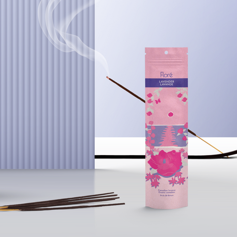 A light pink package of incense sticks that reads Flore Lavender with an image of dark pink flowers framing a blue lake with a pink sun in the sky. The package stands upright next to a burning incense stick against a pale purple background.