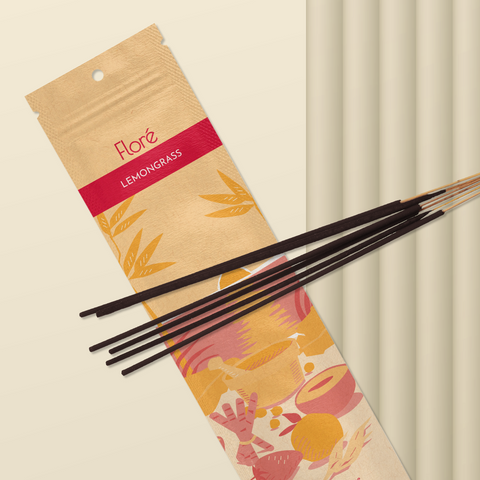 A pale orange package of incense sticks that reads Flore Lemongrass with an image of red and orange fruits, spices and mortar and pestle on a beach at sunset. The package lies with a bundle of incense sticks on a sandy background.