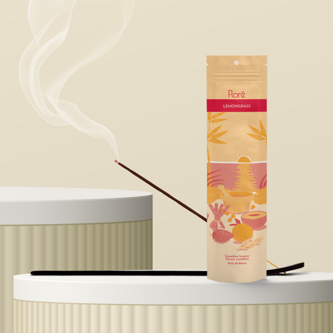 A pale orange package of incense sticks that reads Flore Lemongrass with an image of red and orange fruits, spices and mortar and pestle on a beach at sunset. The package stands upright on a podium next to a burning incense stick against a sandy background.