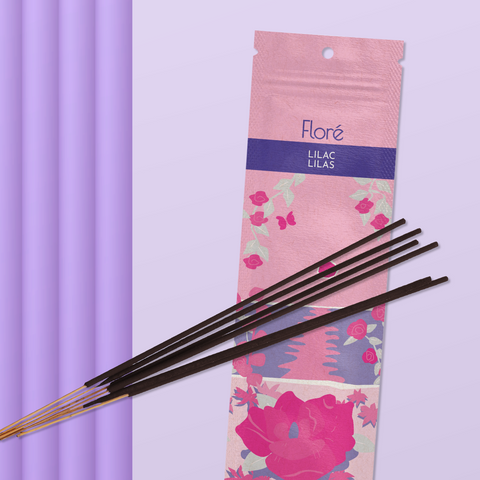 A light pink package of incense sticks that reads Flore Lilac with an image of dark pink flowers framing a blue lake with a pink sun in the sky. The package lies with a bundle of incense sticks on a pale purple background.