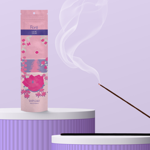 A light pink package of incense sticks that reads Flore Lilac with an image of dark pink flowers framing a blue lake with a pink sun in the sky. The package stands upright on a podium next to a burning incense stick against a pale purple background.