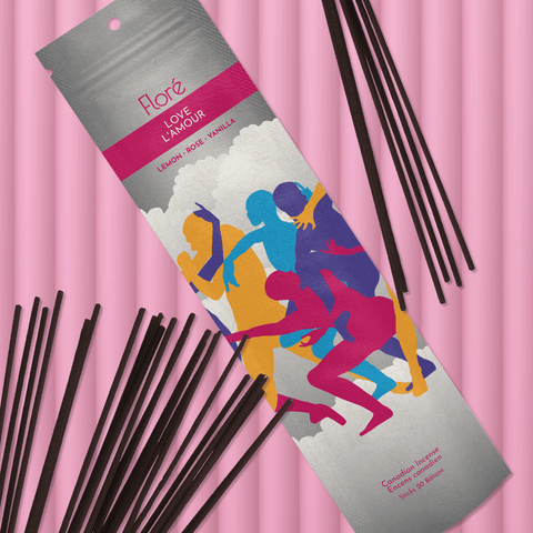 A multi toned grey package of incense sticks that reads Flore Love with an image of multiple bright coloured human silhouettes entangled in various poses against a white and grey cloud background. The package lies with a bundle of incense sticks on a bright pink background.