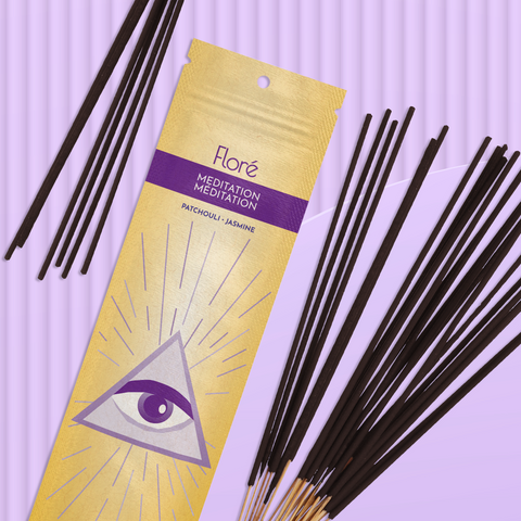 A bright yellow package of incense sticks that reads Flore Meditation with an image of a purple third eye surrounded by a triangle and pink and purple rays of light. The package lies with a bundle of incense sticks on a pale lilac background.