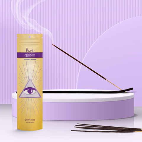 A bright yellow package of incense sticks that reads Flore Meditation with an image of a purple third eye surrounded by a triangle and pink and purple rays of light. The package stands upright on a podium next to a burning incense stick against a pale lilac background. 