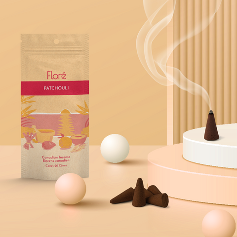 A pale orange package of incense cones that reads Flore Patchouli with an image of red and orange fruits, spices and mortar and pestle on a beach at sunset. The package stands upright on a podium next to a burning incense cone and white orbs against a pale orange background.