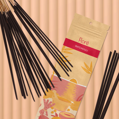 A pale orange package of incense sticks that reads Flore Patchouli with an image of red and orange fruits, spices and mortar and pestle on a beach at sunset. The package lies with bundles of incense sticks on a pale orange background.