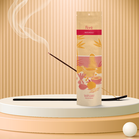 A pale orange package of incense sticks that reads Flore Patchouli with an image of red and orange fruits, spices and mortar and pestle on a beach at sunset. The package stands upright on a podium next to a burning incense stick and white orbs against a pale orange background.