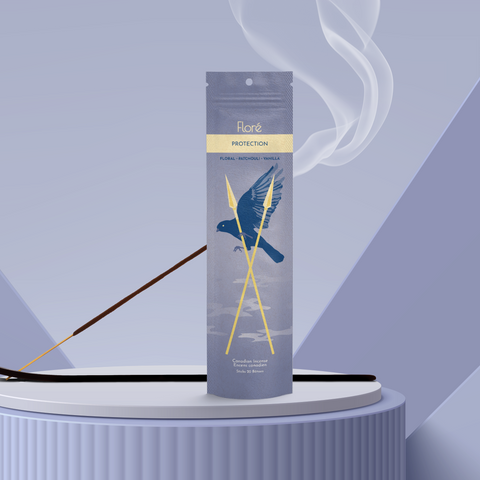 A smoky purple package of incense sticks that reads Flore Protection with an image of a flying crow behind crossed golden spears. The package stands upright on a podium next to a burning incense stick against a periwinkle background.