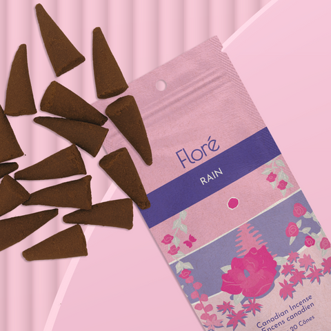 A light pink package of incense cones that reads Flore Rain with an image of dark pink flowers framing a blue lake with a pink sun in the sky. The package lies with a bunch of incense cones on a pale pink background.