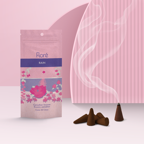 A light pink package of incense cones that reads Flore Rain with an image of dark pink flowers framing a blue lake with a pink sun in the sky. The package stands upright on a podium next to a burning incense cone against a pale pink background.