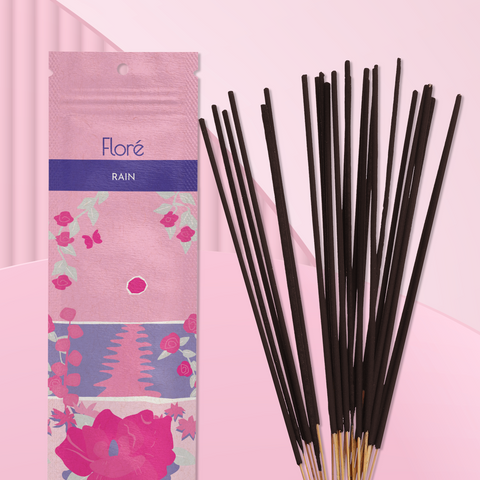 A light pink package of incense sticks that reads Flore Rain with an image of dark pink flowers framing a blue lake with a pink sun in the sky. The package lies with a bundle of incense sticks on a pale pink background.