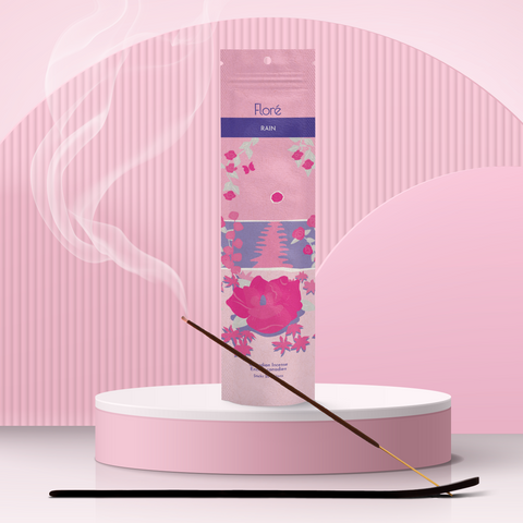 A light pink package of incense sticks that reads Flore Rain with an image of dark pink flowers framing a blue lake with a pink sun in the sky. The package stands upright on a podium next to a burning incense stick against a pale pink background.
