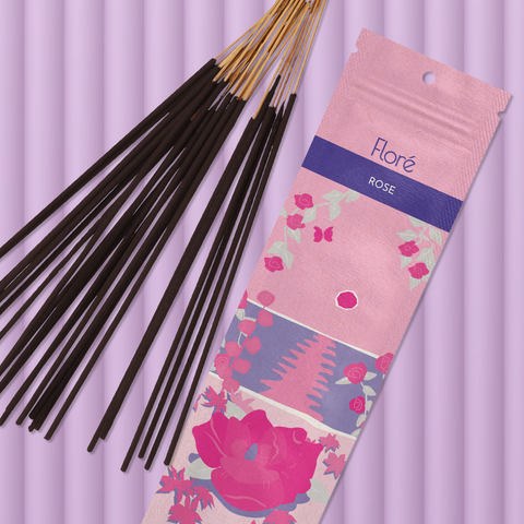 A light pink package of incense sticks that reads Flore Rose with an image of dark pink flowers framing a blue lake with a pink sun in the sky. The package lies with a bundle of incense sticks on a pale purple background.