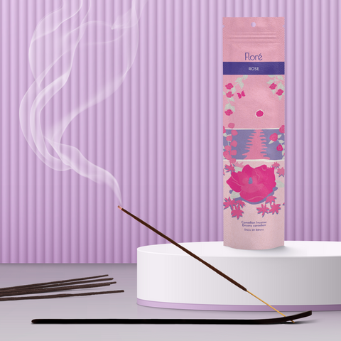 A light pink package of incense sticks that reads Flore Rose with an image of dark pink flowers framing a blue lake with a pink sun in the sky. The package stands upright on a podium next to a burning incense stick against a pale purple background.