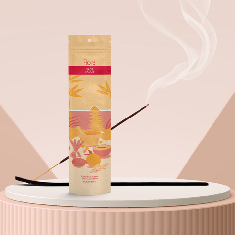 A pale orange package of incense sticks that reads Flore Sage with an image of red and orange fruits, spices and mortar and pestle on a beach at sunset. The package stands upright on a podium next to a burning incense stick against a rosy taupe background.