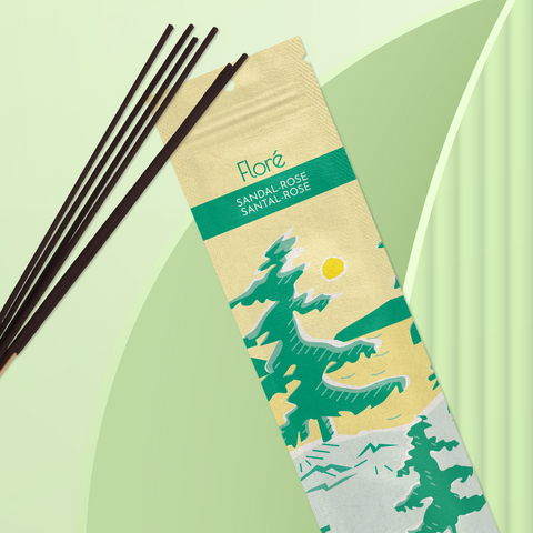 A pale yellow package of incense sticks that reads Flore Sandal-Rose with an image of pine trees growing on the shore of a golden lake with a yellow sun in the sky. The package lies with a bundle of incense sticks on a mint green background.