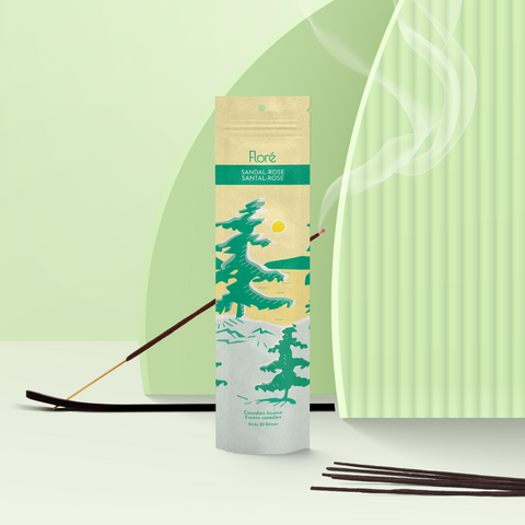 A pale yellow package of incense sticks that reads Flore Sandal-Rose with an image of pine trees growing on the shore of a golden lake with a yellow sun in the sky. The package stands upright next to a burning incense stick against a mint green background.