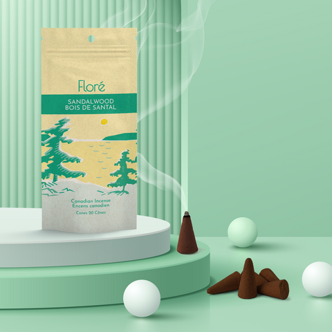 A pale yellow package of incense cones that reads Flore Sandalwood with an image of pine trees growing on the shore of a golden lake with a yellow sun in the sky. The package stands upright next to a burning incense cone and white orbs against a pale green background.
