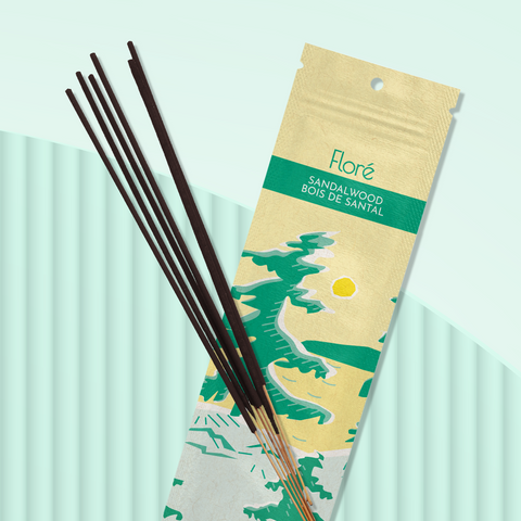 A pale yellow package of incense sticks that reads Flore Sandalwood with an image of pine trees growing on the shore of a golden lake with a yellow sun in the sky. The package lies with a bundle of incense sticks on a pale green background.