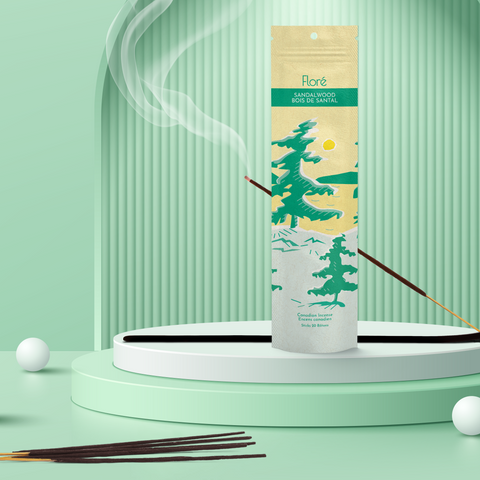 A pale yellow package of incense sticks that reads Flore Sandalwood with an image of pine trees growing on the shore of a golden lake with a yellow sun in the sky. The package stands upright next to a burning incense stick and white orbs against a pale green background.