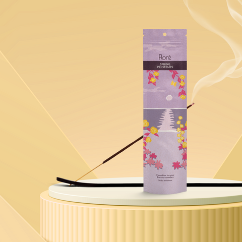 A pale purple package of incense sticks that reads Flore Spring with an image of yellow flowers framing a purple lake with a grey sun in the sky. The package stands upright on a podium next to a burning incense stick against a pale golden background.
