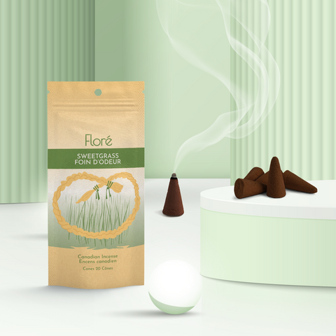 A pale gold and green package of incense cones that reads Flore Sweetgrass with an image of a circular golden sweetgrass braid in front of tall thin green grass. The package stands upright on a podium next to a burning incense cone and white orbs against a pale mossy green background.