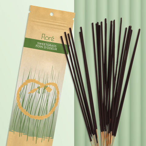 A pale gold and green package of incense sticks that reads Flore Sweetgrass with an image of a circular golden sweetgrass braid in front of tall thin green grass. The package lies with a bundle of incense sticks on a pale mossy green background.