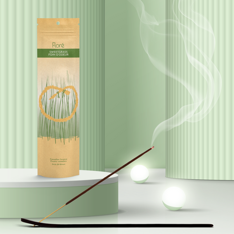 A pale gold and green package of incense sticks that reads Flore Sweetgrass with an image of a circular golden sweetgrass braid in front of tall thin green grass. The package stands upright on a podium next to a burning incense stick and white orbs against a pale mossy green background.