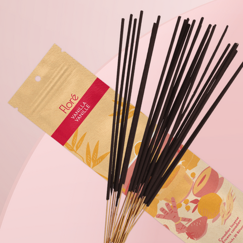 A pale orange package of incense sticks that reads Flore Vanilla with an image of red and orange fruits, spices and mortar and pestle on a beach at sunset. The package lies with a bundle of incense sticks on a rosy peach background.