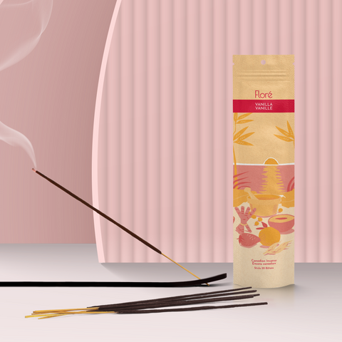 A pale orange package of incense sticks that reads Flore Vanilla with an image of red and orange fruits, spices and mortar and pestle on a beach at sunset. The package stands upright next to a burning incense stick against a rosy peach background.