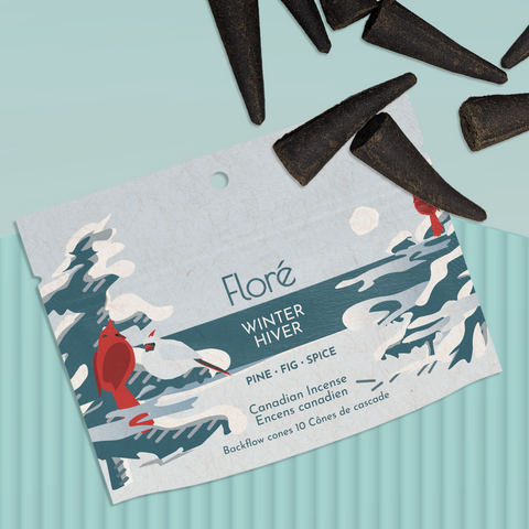 A pale cool grey package of backflow incense cones that reads Flore Winter with an image of two male cardinals and one female cardinal sitting on the branches of dark blue-grey pine trees on the shore of a blue lake with a white sun in the sky. The package lies with a bunch of incense cones on a pale teal background.