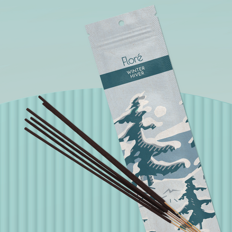 A pale cool grey package of incense sticks that reads Flore Winter with an image of dark blue-grey pine trees growing on the shore of a blue lake with a white sun in the sky. The package lies with a bundle of incense sticks on a pale teal background.