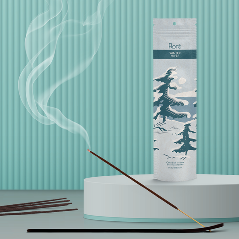 A pale cool grey package of incense sticks that reads Flore Winter with an image of dark blue-grey pine trees growing on the shore of a blue lake with a white sun in the sky. The package stands upright next to a burning incense stick against a pale teal background.