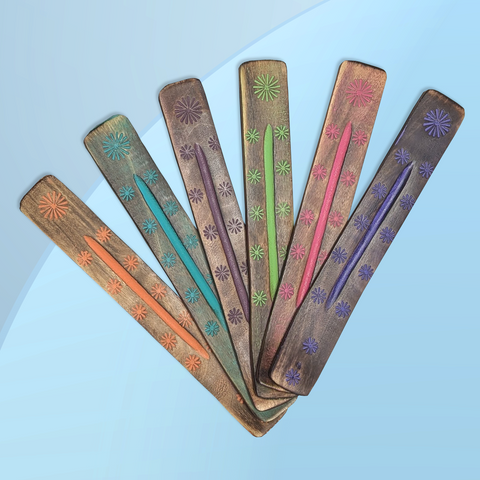 Six long flat wooden incense burners each featuring a different colour floral design laying side by side diagonally on a blue background. From left to right the colours of the incense burners are peach, teal, merlot, lime green, pink, purple.