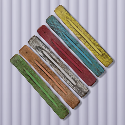 Six long flat wooden incense burners each a different colour laying side by side diagonally on a pink background. From left to right the colours of the incense burners are a faded green, peach, white, red, blue, yellow.