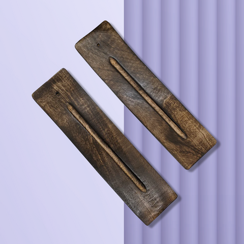 Two wide flat dark wooden incense burners each with a long single groove carved lengthwise, laying side by side diagonally on a purple background.
